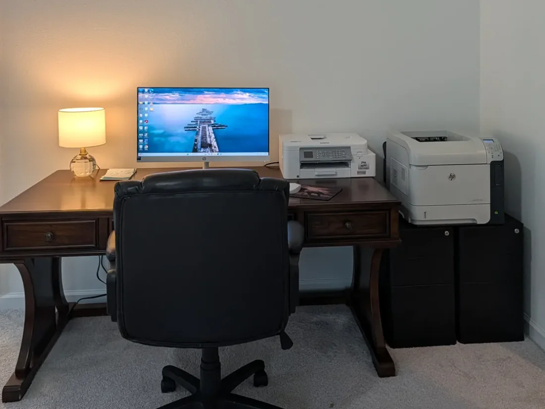 Home office desk with an all-in-one computer, a desk lamp and two printers.